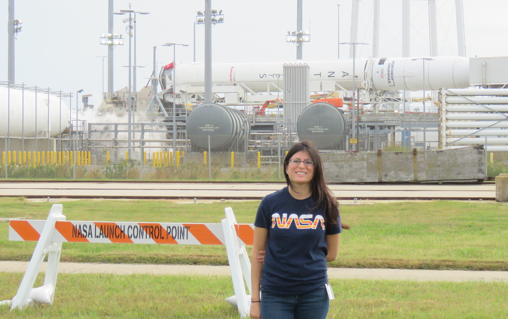 A person wearing jeans and a navy blue shirt with "NASA" written in bold orange and white letters on it. The person is outside during the day on a launch pad.